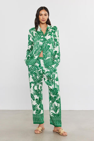 A woman wearing a green and white palm print pantsuit from Velvet by Graham & Spencer, with a matching crop top, standing against a plain backdrop, paired with beige sandals.