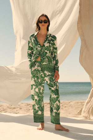 A woman in a green Velvet by Graham & Spencer AYLA button-up shirt stands on a beach, flanked by billowing white fabrics, with sunglasses on.