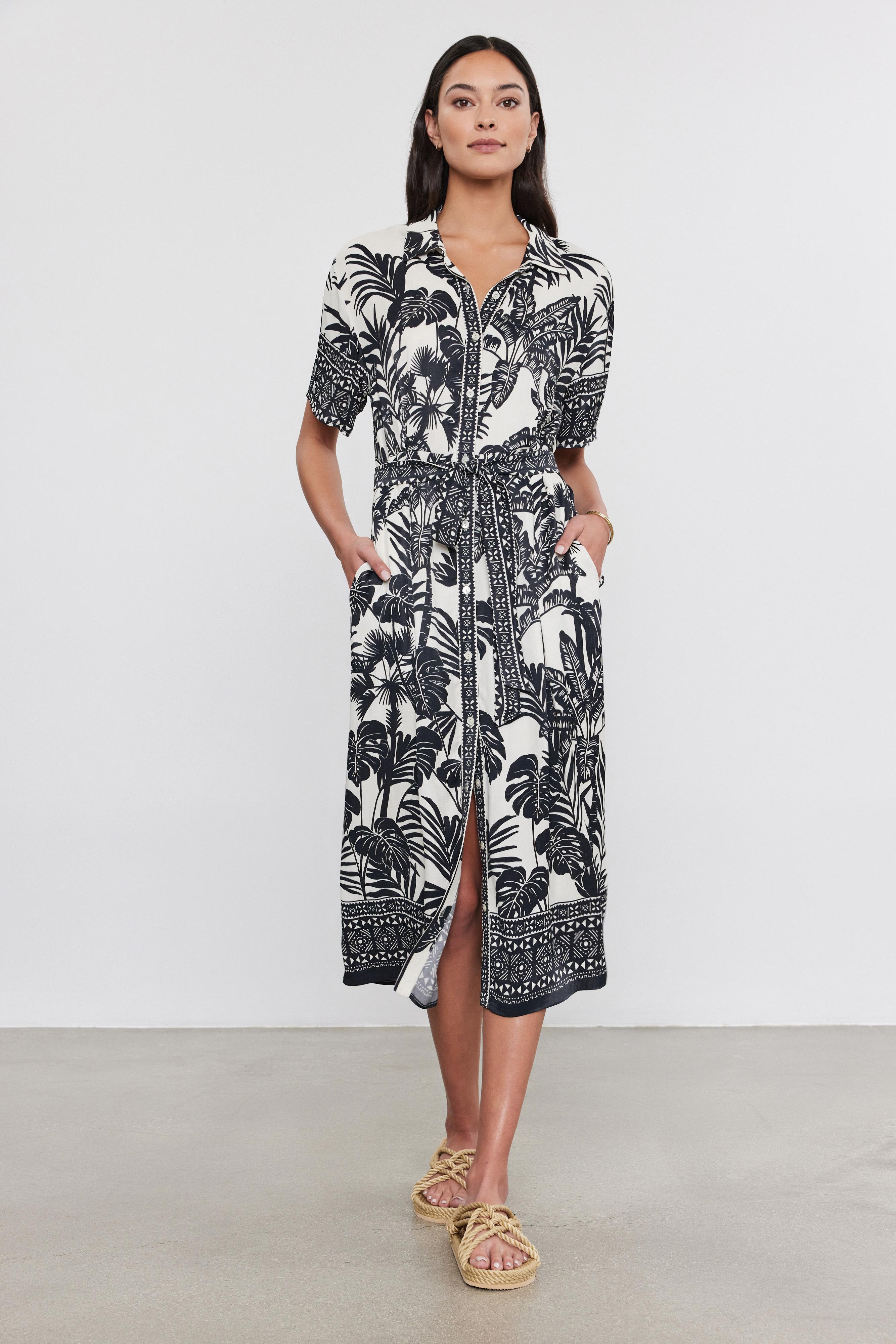   A woman stands in a studio wearing a black and white palm print Freya Dress by Velvet by Graham & Spencer with a front zipper and sandals. 