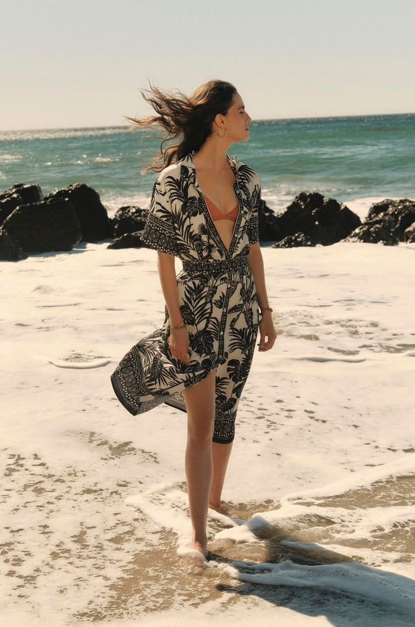 Woman in a tropical Freya dress by Velvet by Graham & Spencer standing on a sandy beach with waves touching her feet, looking out to sea, hair blowing in the wind.