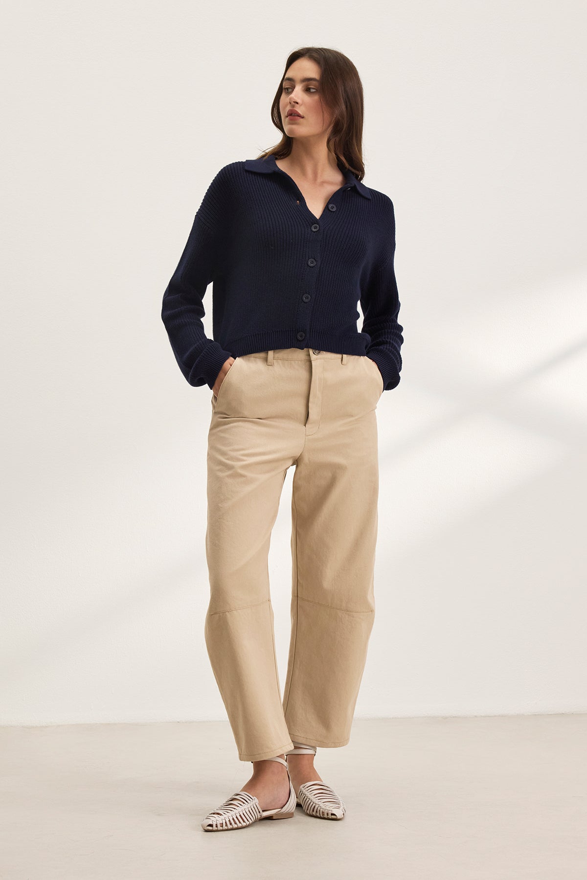   A person stands against a plain background wearing a dark blue button-up sweater, Velvet by Graham & Spencer's ALEXIS PANT in cropped beige cotton twill, and white woven shoes. 