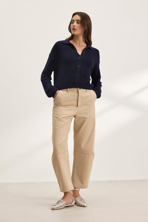 A person stands against a plain background wearing a dark blue button-up sweater, Velvet by Graham & Spencer's ALEXIS PANT in cropped beige cotton twill, and white woven shoes.