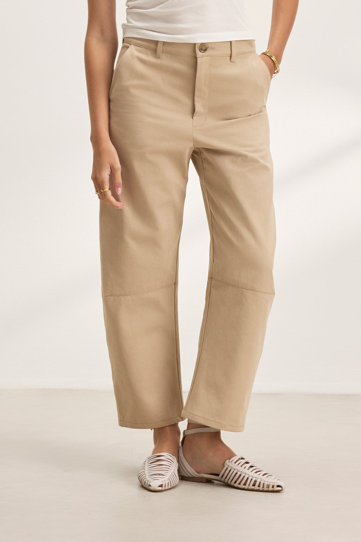   A person stands against a plain background wearing the ALEXIS PANT by Velvet by Graham & Spencer, a white top, and white woven sandals, with one hand casually tucked in their pocket. 