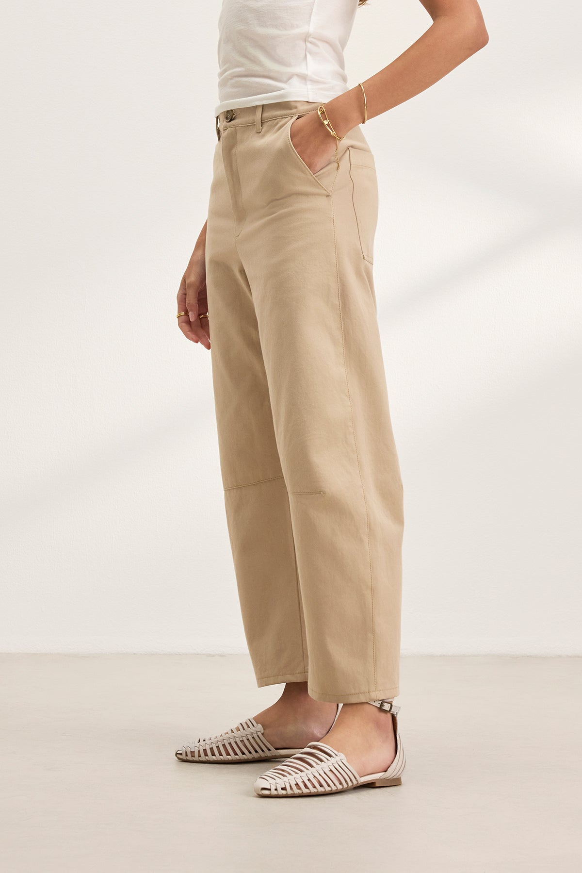   A person confidently stands against a plain background wearing the ALEXIS PANT, featuring a white top and beige cropped cotton twill pants with knee darting from Velvet by Graham & Spencer, paired with white woven sandals and one hand in their pocket. 