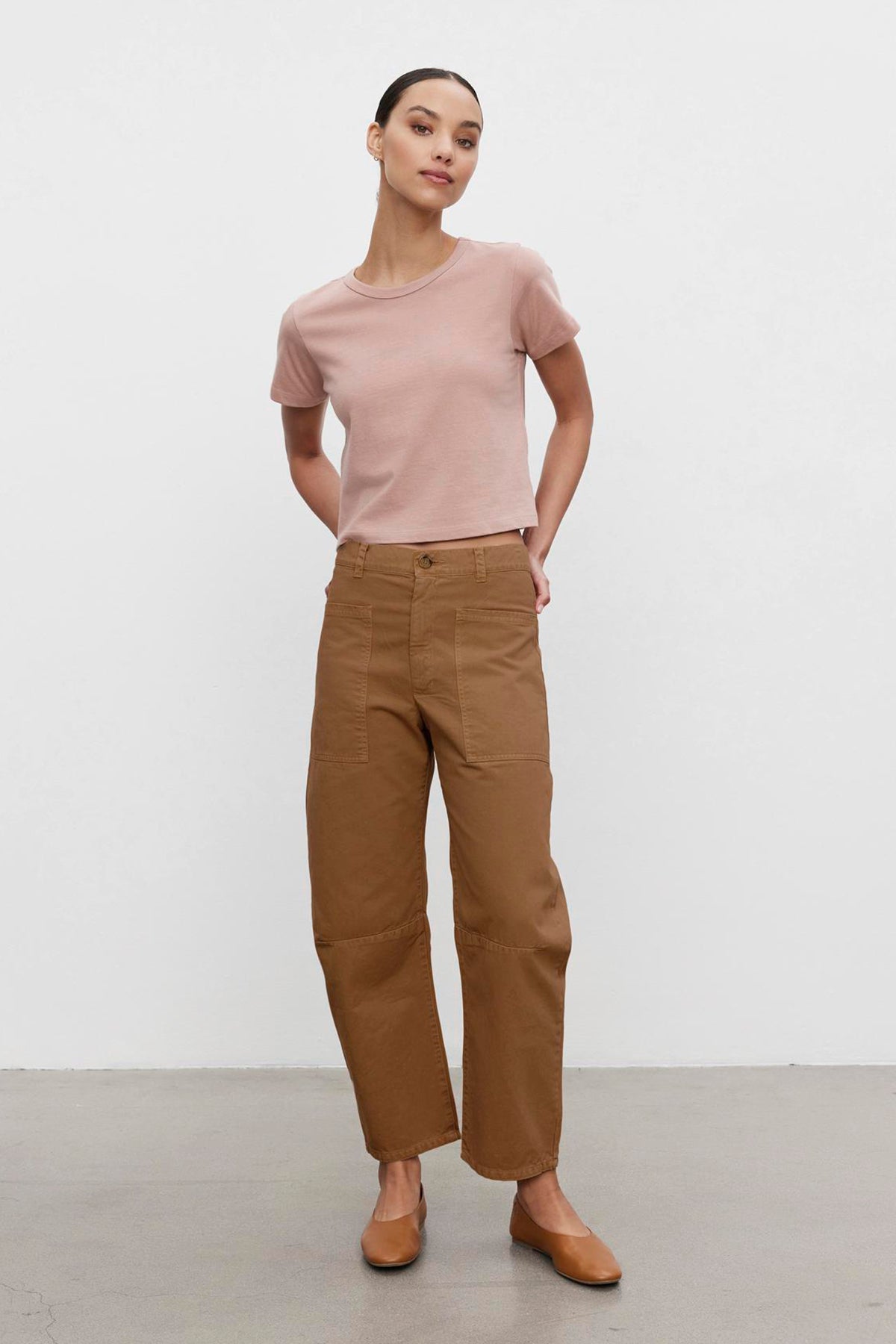 A woman wearing a light pink T-shirt, BRYLIE SANDED TWILL UTILITY PANT from Velvet by Graham & Spencer, and brown shoes stands against a plain white background with her hands in her pockets.-37676285558977