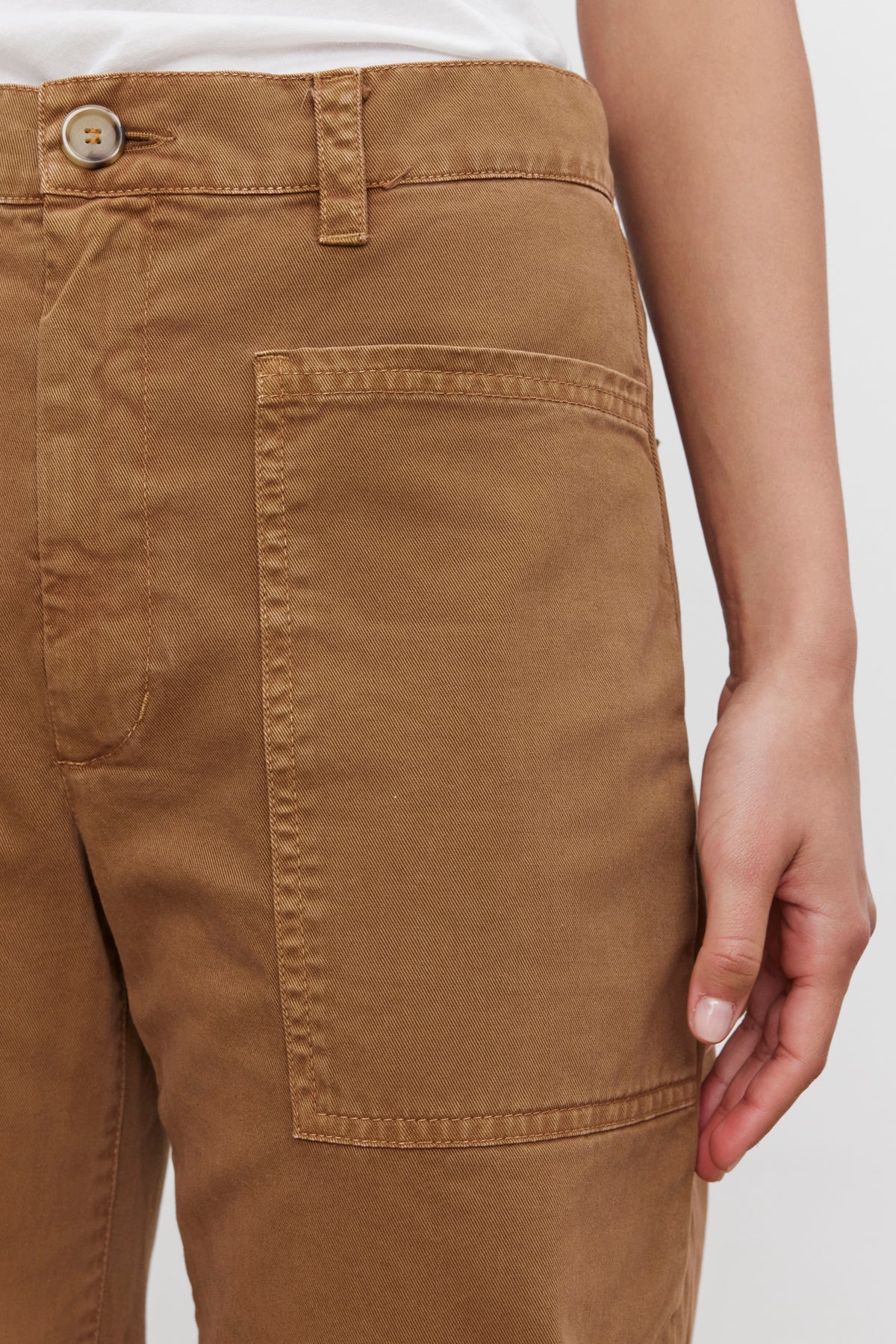   Close-up of a person wearing the BRYLIE SANDED TWILL UTILITY PANT by Velvet by Graham & Spencer, featuring a buttoned waistband and large front pocket. The casual brown cotton trouser showcases a utilitarian-inspired design. The person's hand is resting by their side, with the background plain and out of focus. 