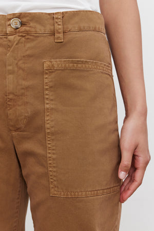 Close-up of a person wearing the BRYLIE SANDED TWILL UTILITY PANT by Velvet by Graham & Spencer, featuring a buttoned waistband and large front pocket. The casual brown cotton trouser showcases a utilitarian-inspired design. The person's hand is resting by their side, with the background plain and out of focus.