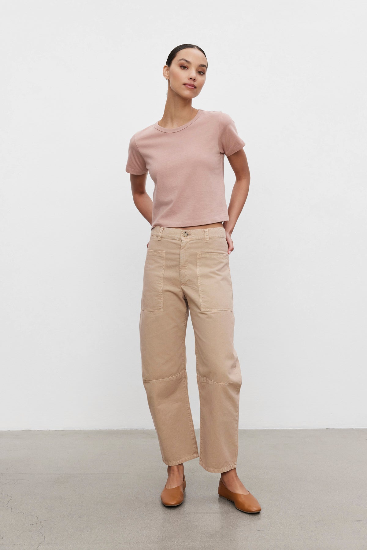   A person wearing the TINSLEY TEE from Velvet by Graham & Spencer, which is a light pink cropped tee with a vintage feel, beige pants, and brown shoes stands against a plain white background with their hands behind their back. 