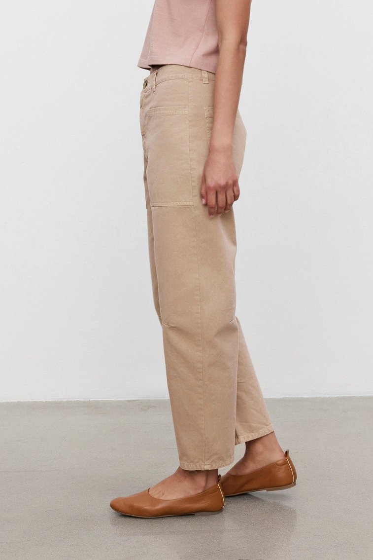   A person with tied-back hair, wearing a pink t-shirt, BRYLIE SANDED TWILL UTILITY PANT by Velvet by Graham & Spencer, and brown flats, standing in profile against a plain white background. 