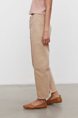 A person with tied-back hair, wearing a pink t-shirt, BRYLIE SANDED TWILL UTILITY PANT by Velvet by Graham & Spencer, and brown flats, standing in profile against a plain white background.