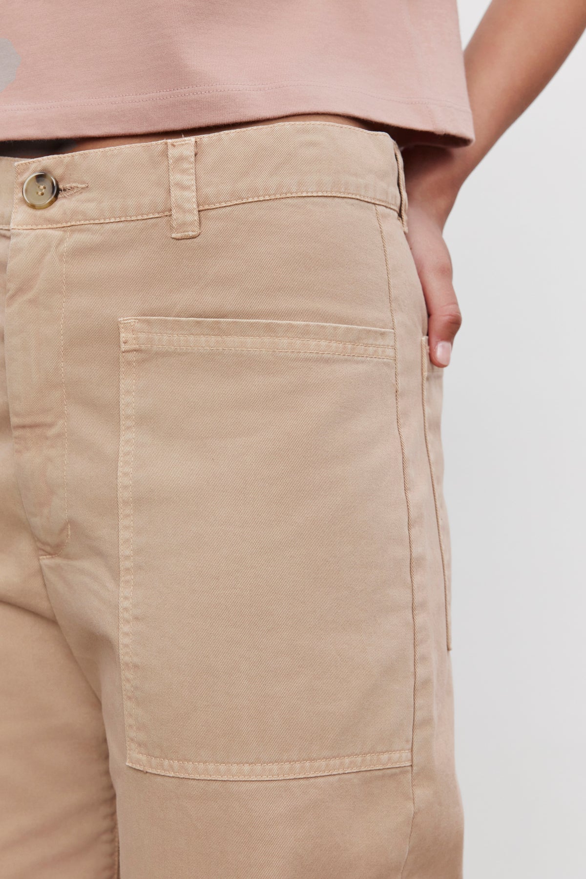   Close-up of a person wearing the BRYLIE SANDED TWILL UTILITY PANT by Velvet by Graham & Spencer. The beige cotton twill pants feature large patch pockets, with the person's hand resting inside one pocket, and a partial view of a pink shirt in the background. 