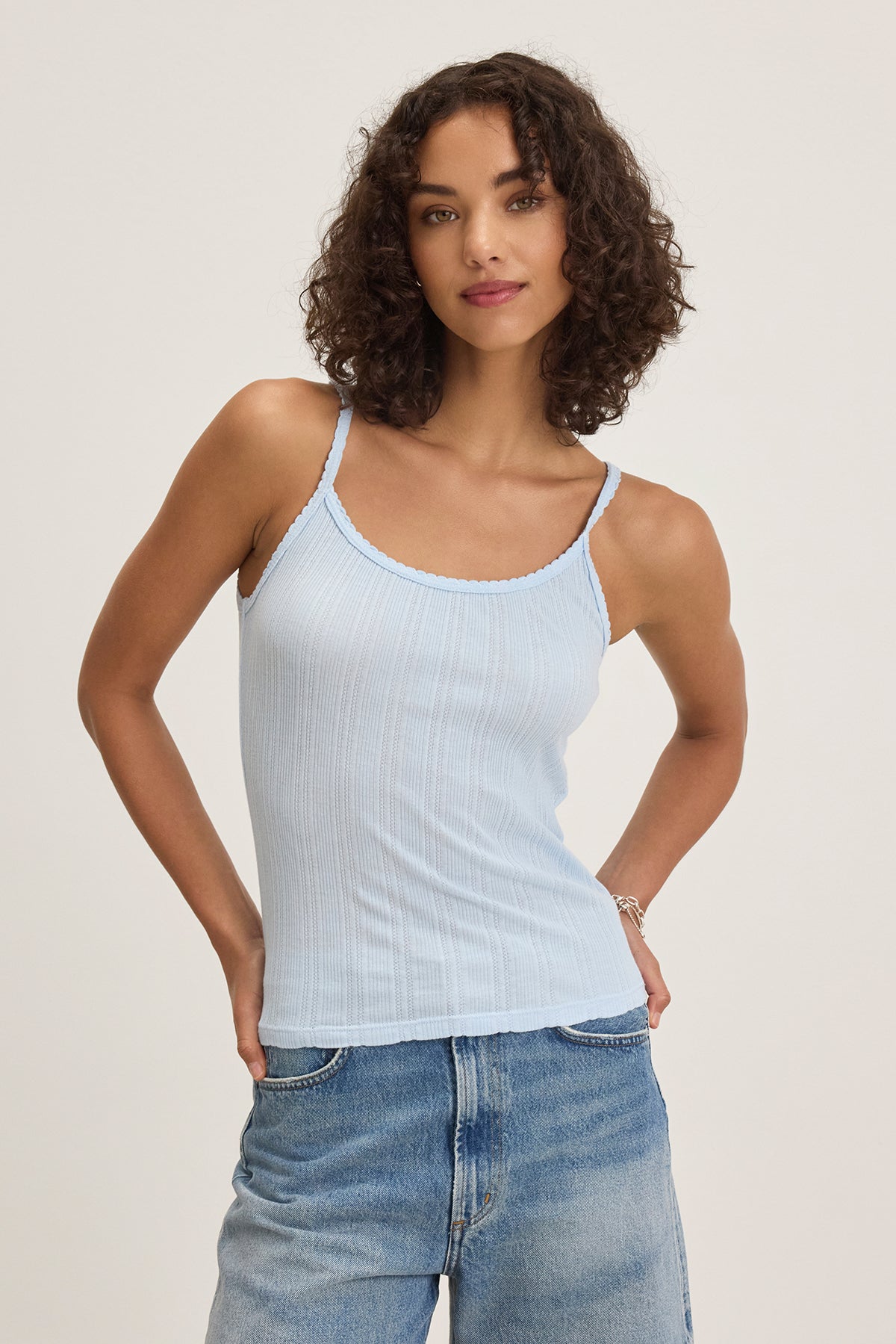   A person with curly hair models the INIKU TANK TOP by Velvet by Graham & Spencer, a light blue sleeveless top with scalloped edges, paired with jeans against a plain background. 