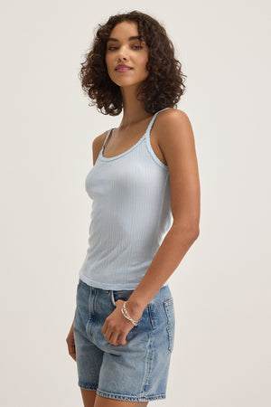 A person in the INIKU TANK TOP by Velvet by Graham & Spencer and denim shorts, standing and smiling against a plain background.