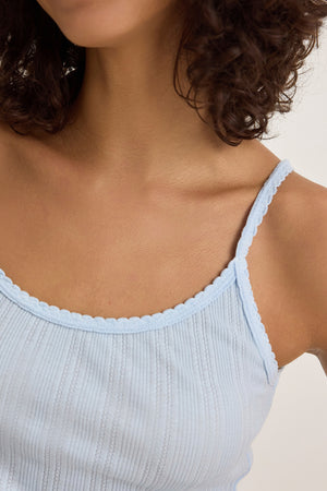 A person with curly hair wears the INIKU TANK TOP from Velvet by Graham & Spencer, in a light blue pointelle ribbed cotton modal blend with scalloped edges, captured from neck to chest.