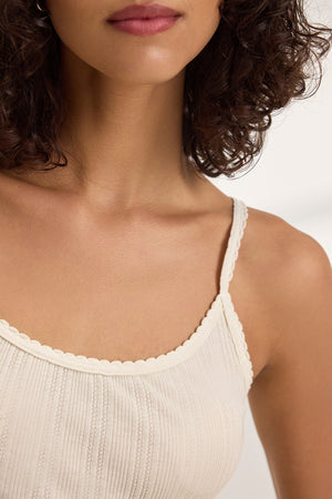 A person with curly hair is wearing the INIKU TANK TOP by Velvet by Graham & Spencer, a cream-colored, cotton modal blend with a subtle pointelle ribbed texture.