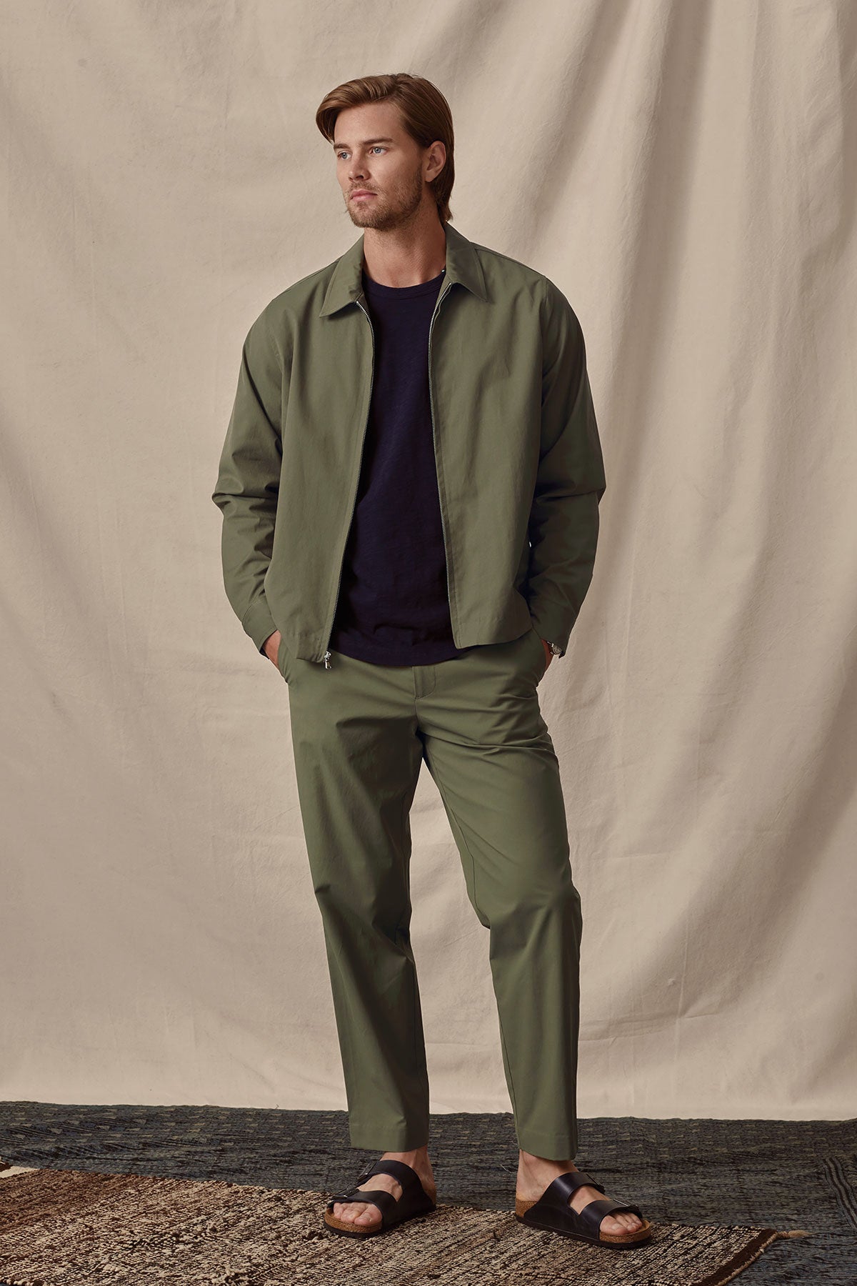   A man in a casual olive green jacket and STING POPLIN PANT trousers with a tailored style, made from a cotton blend, posing against a beige background by Velvet by Graham & Spencer. 