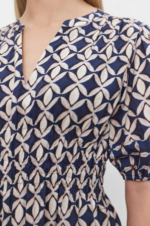 Close-up of a person wearing the JOEY DRESS by Velvet by Graham & Spencer, featuring a patterned blue, white, and beige printed cotton cambric fabric with a deep v-neckline and gathered waist detail.