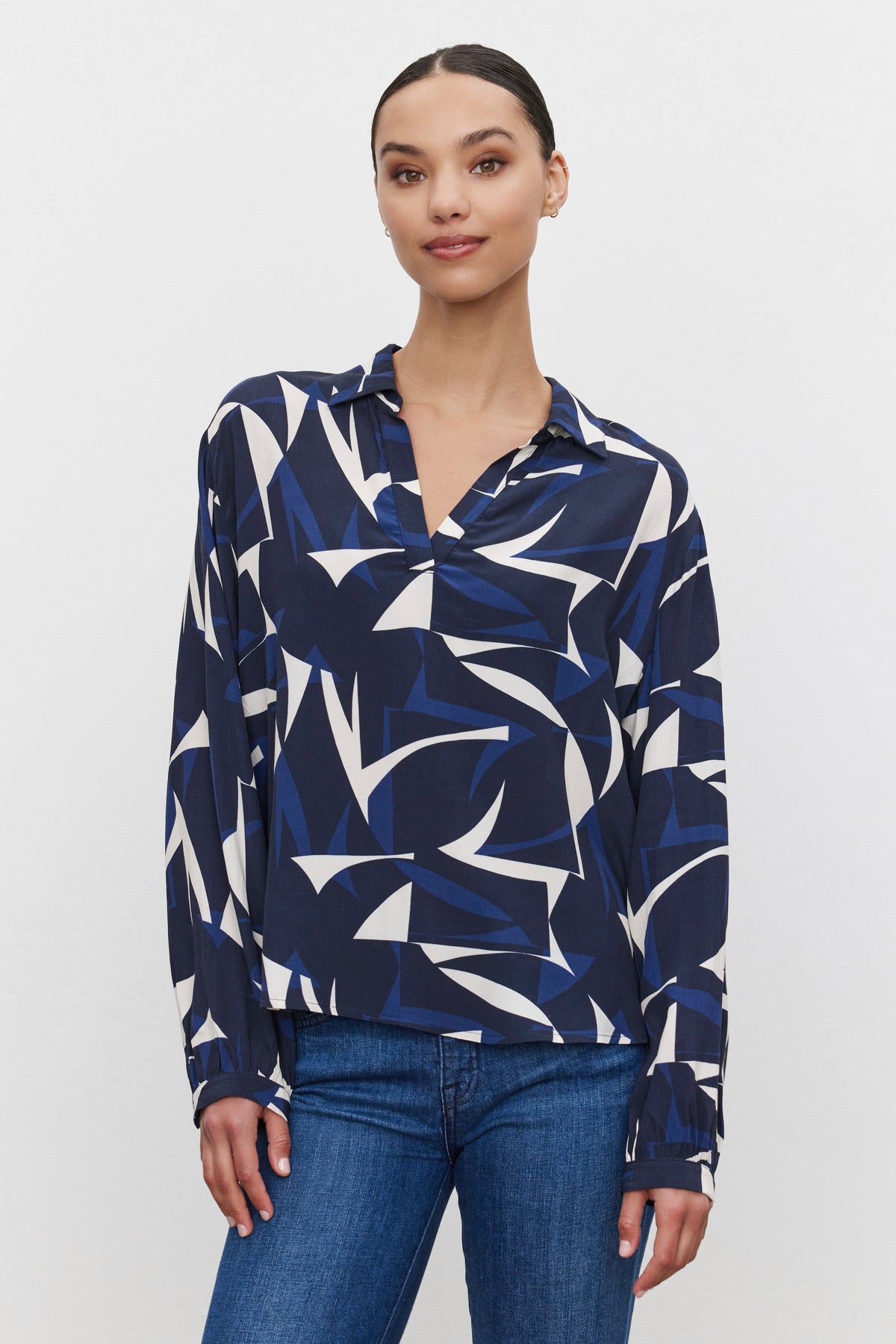   A person stands against a plain background, wearing the Velvet by Graham & Spencer CARAH TOP, a long-sleeve, split-neck blouse in blue, white, and black geometric shapes made from printed royal crepe fabric, paired with blue jeans. 