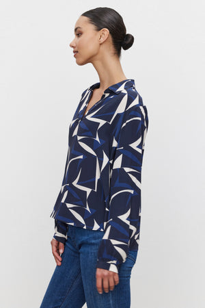 A person wearing the CARAH TOP by Velvet by Graham & Spencer, a blue and white split neck blouse with long sleeves, paired with blue jeans stands against a plain background, shown in a side profile.