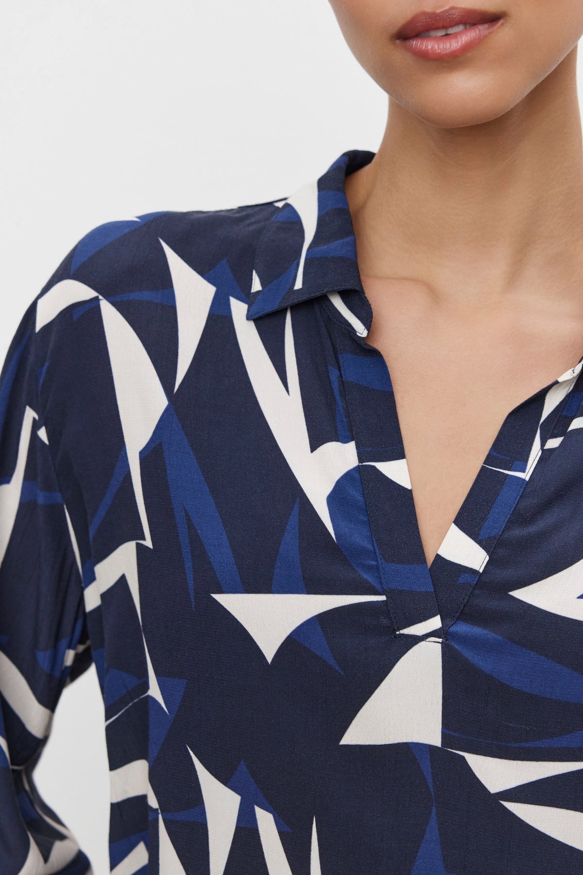   Close-up of a person wearing the CARAH TOP from Velvet by Graham & Spencer, showcasing its dark blue fabric adorned with an abstract white and light blue geometric pattern. The design features a split neckline and collar, with textured fabric that adds depth. The face is partially visible in the image. 