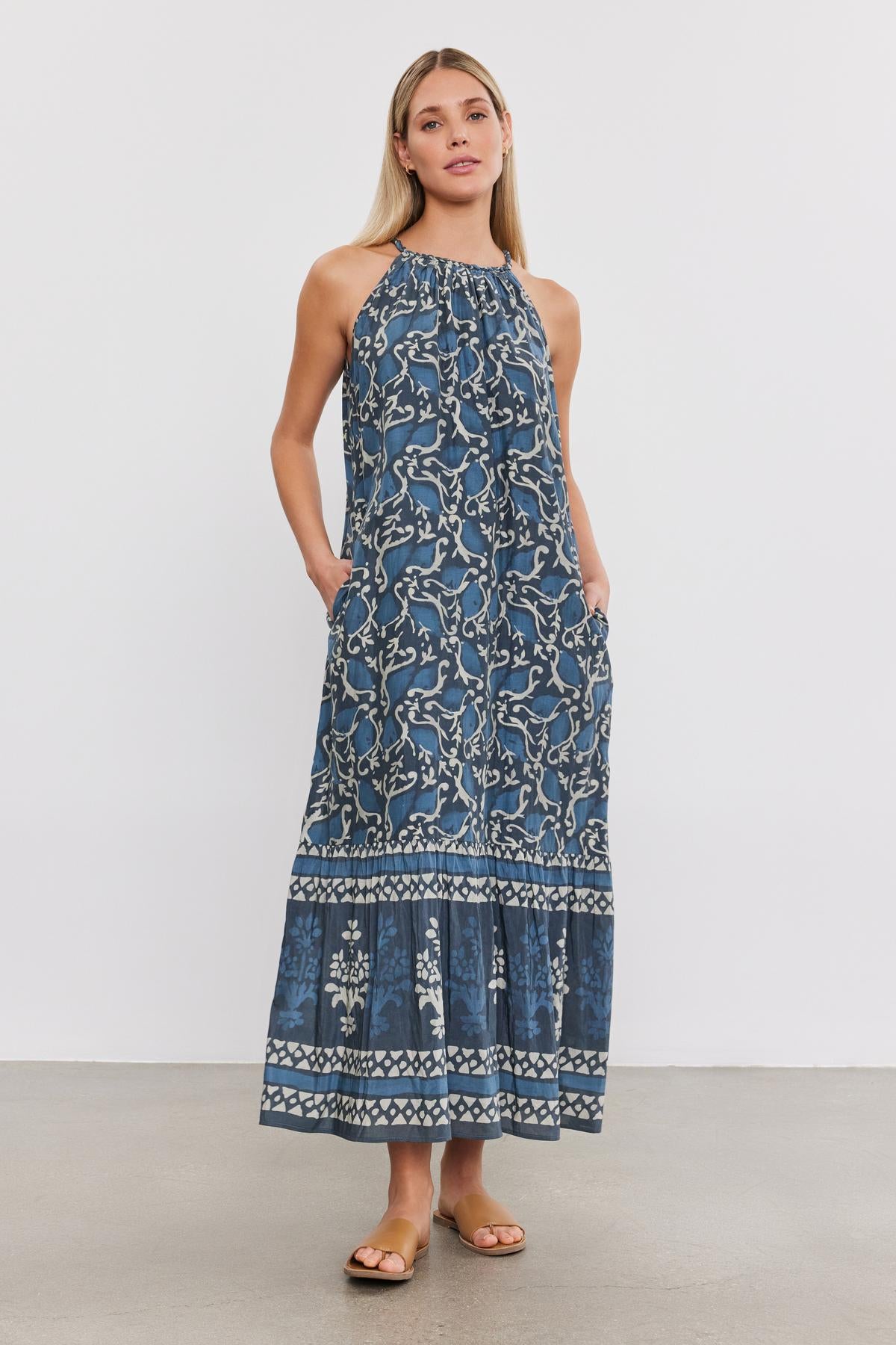 A woman stands in a blue, patterned, sleeveless, ankle-length GHITA DRESS with pockets by Velvet by Graham & Spencer. She is wearing simple sandals against a plain white background.-37934969618625