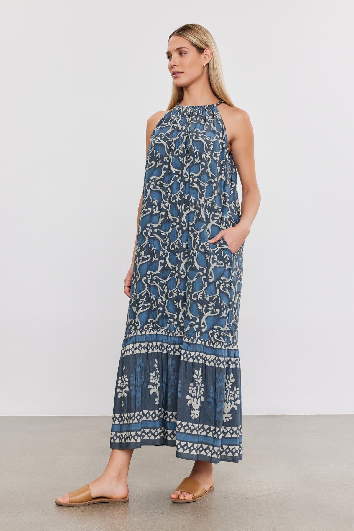   Person standing in the GHITA DRESS by Velvet by Graham & Spencer, a blue sleeveless summer maxi dress with a floral pattern, hands in pockets, and wearing brown sandals. 