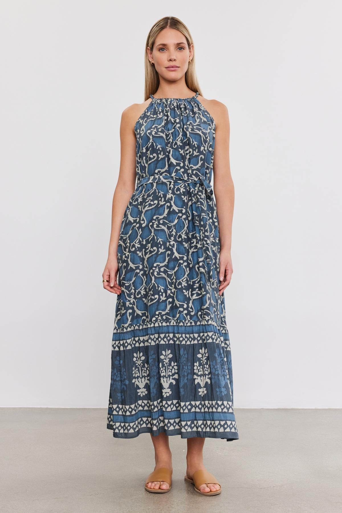   A woman with long hair stands against a white background, wearing the GHITA DRESS, a sleeveless blue silk cotton voile dress adorned with a white floral pattern by Velvet by Graham & Spencer, and brown sandals. 