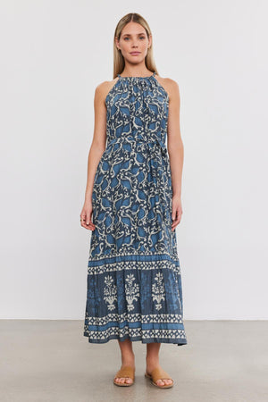 A woman with long hair stands against a white background, wearing the GHITA DRESS, a sleeveless blue silk cotton voile dress adorned with a white floral pattern by Velvet by Graham & Spencer, and brown sandals.