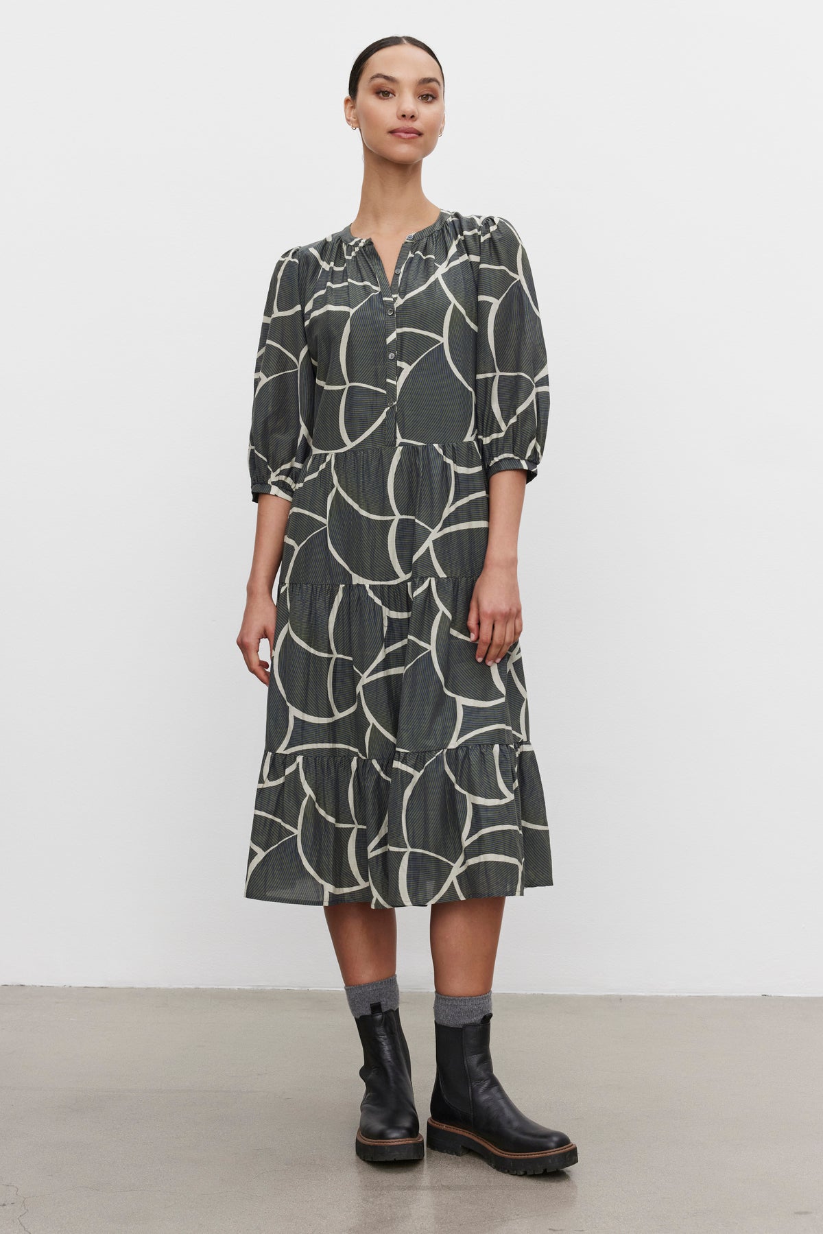   A person stands against a white background wearing the JOHANNA PRINTED SILK COTTON VOILE DRESS by Velvet by Graham & Spencer, featuring long sleeves and a midi-length skirt, paired with black ankle boots. 