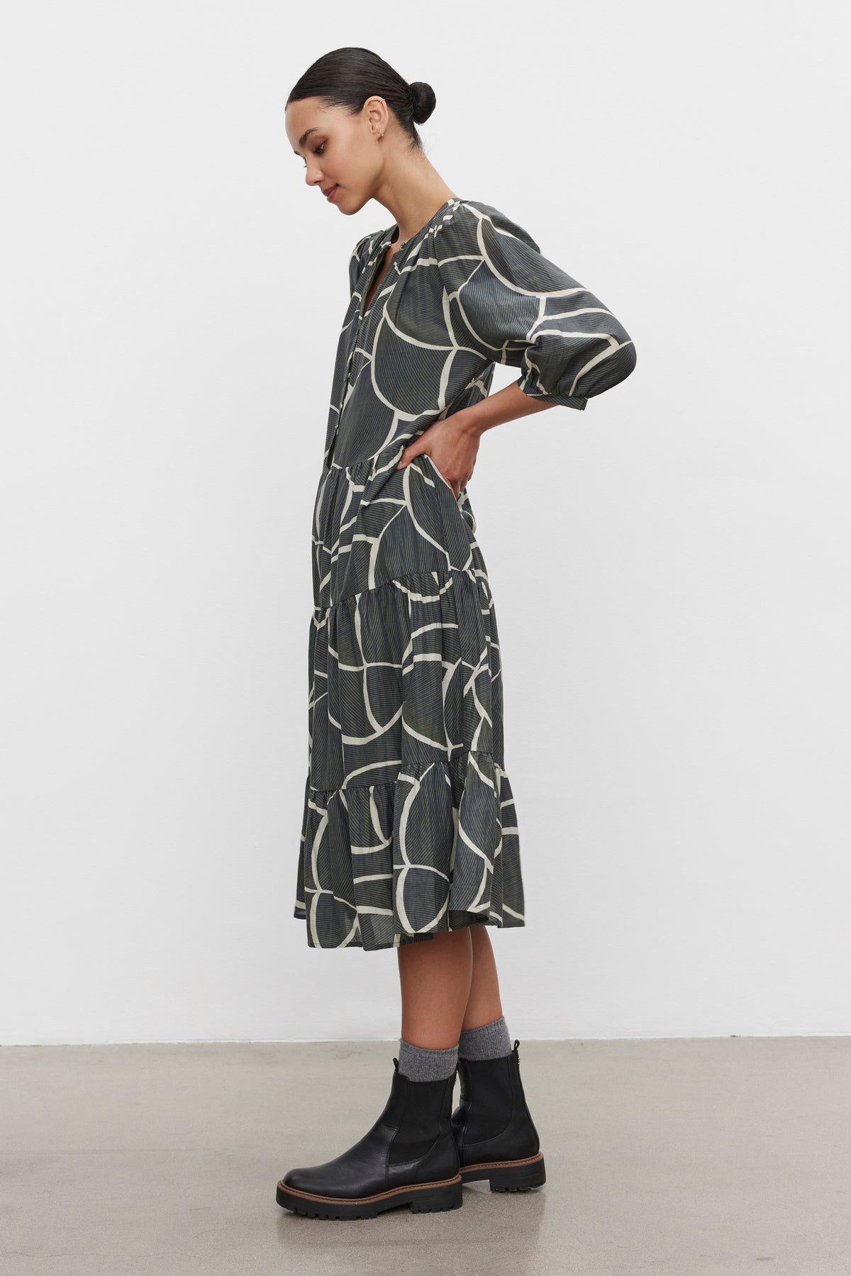   A person stands in profile wearing the JOHANNA PRINTED SILK COTTON VOILE DRESS by Velvet by Graham & Spencer, featuring a bold geometric print. They have short hair tied back and are accessorized with black ankle boots. 