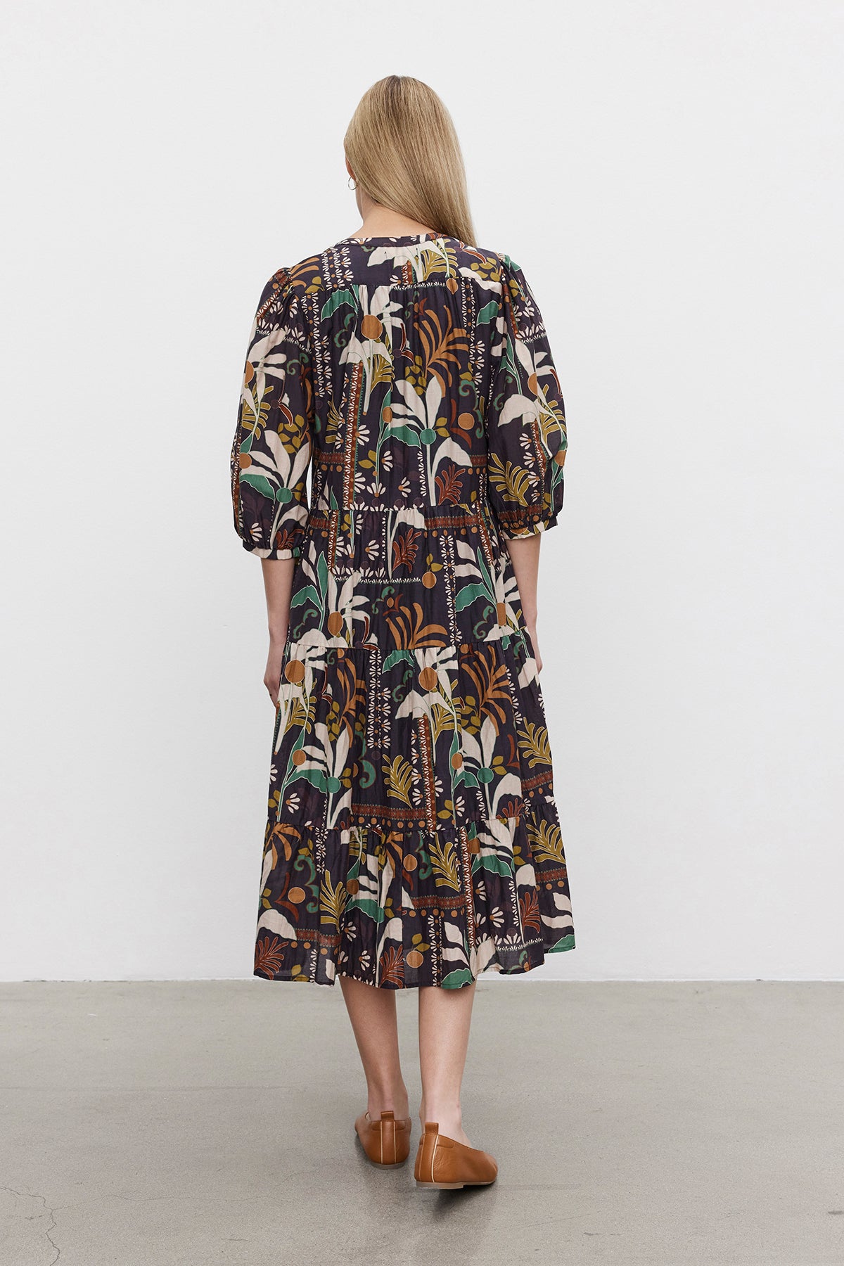   Rear view of a person with long blonde hair wearing the JOHANNA PRINTED SILK COTTON VOILE DRESS by Velvet by Graham & Spencer. The dress features a dark, patterned, long-sleeve design with a button front placket and a three-tiered skirt. They are also wearing brown shoes. The background is plain white. 