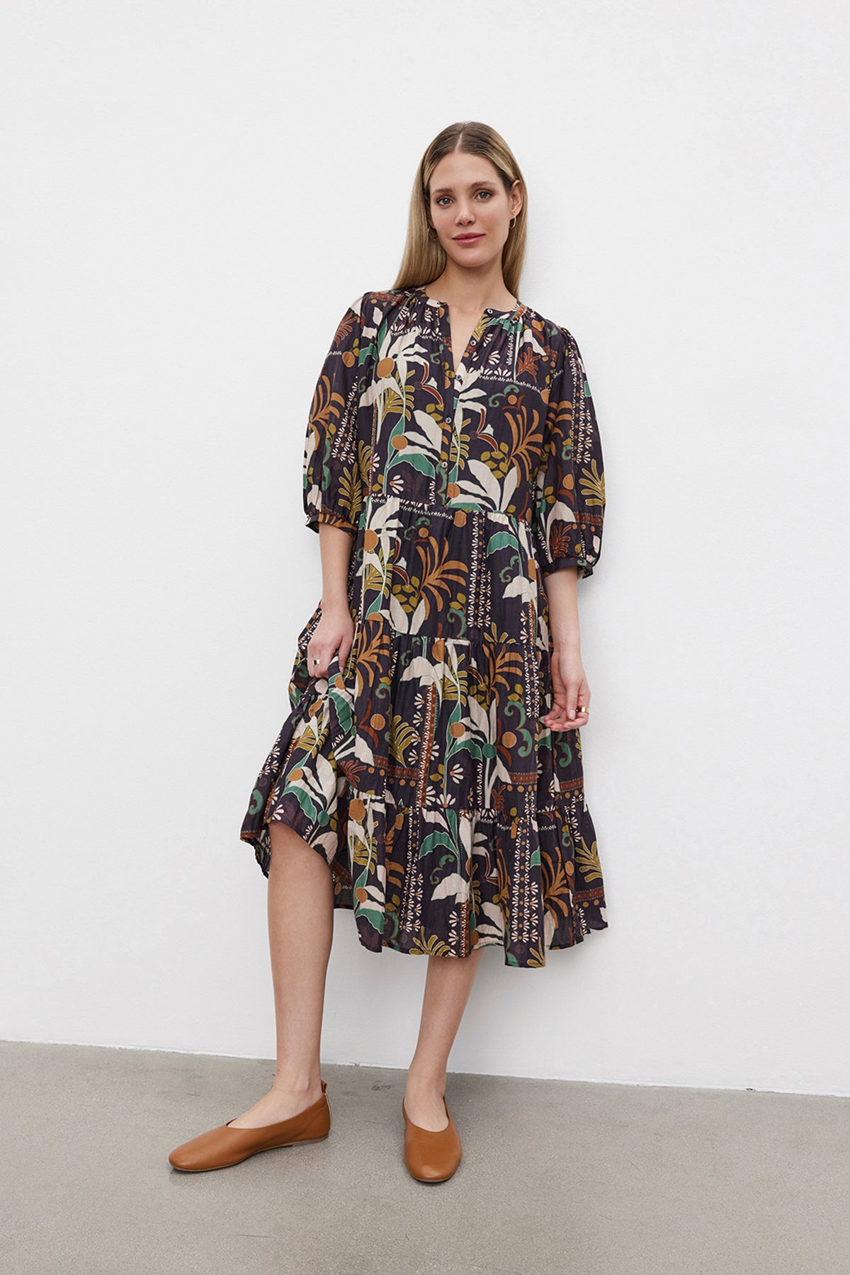   A woman stands against a white wall, wearing the JOHANNA PRINTED SILK COTTON VOILE DRESS by Velvet by Graham & Spencer with puff sleeves and brown flat shoes. The dark floral-patterned dress features a button front placket and is made of luxurious silk cotton voile. 