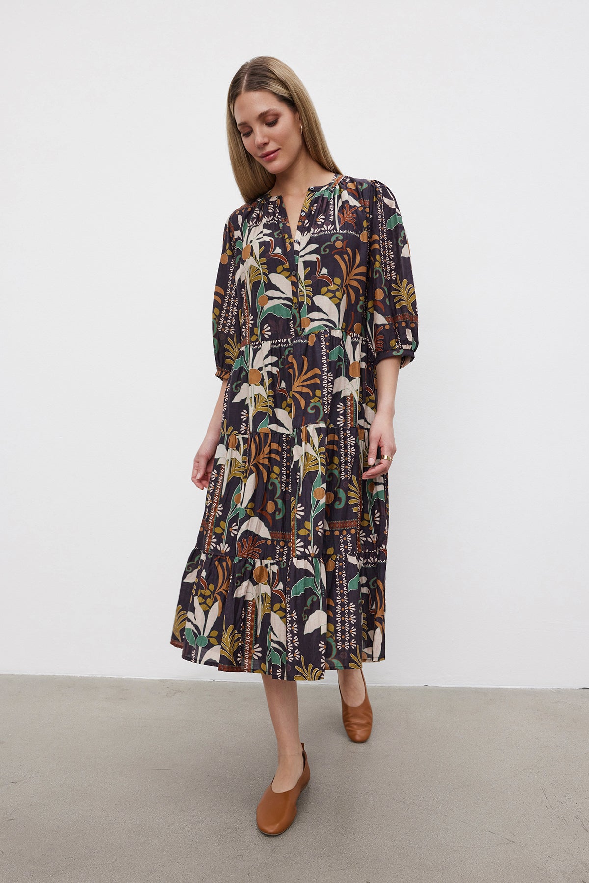   A woman stands against a plain background wearing the JOHANNA PRINTED SILK COTTON VOILE DRESS from Velvet by Graham & Spencer, featuring a knee-length, floral pattern, three-quarter sleeves, and a button front placket. She pairs the dress with brown shoes. 