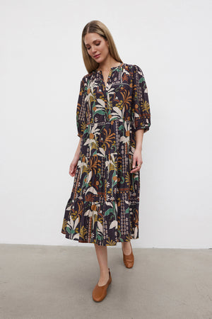 A woman stands against a plain background wearing the JOHANNA PRINTED SILK COTTON VOILE DRESS from Velvet by Graham & Spencer, featuring a knee-length, floral pattern, three-quarter sleeves, and a button front placket. She pairs the dress with brown shoes.