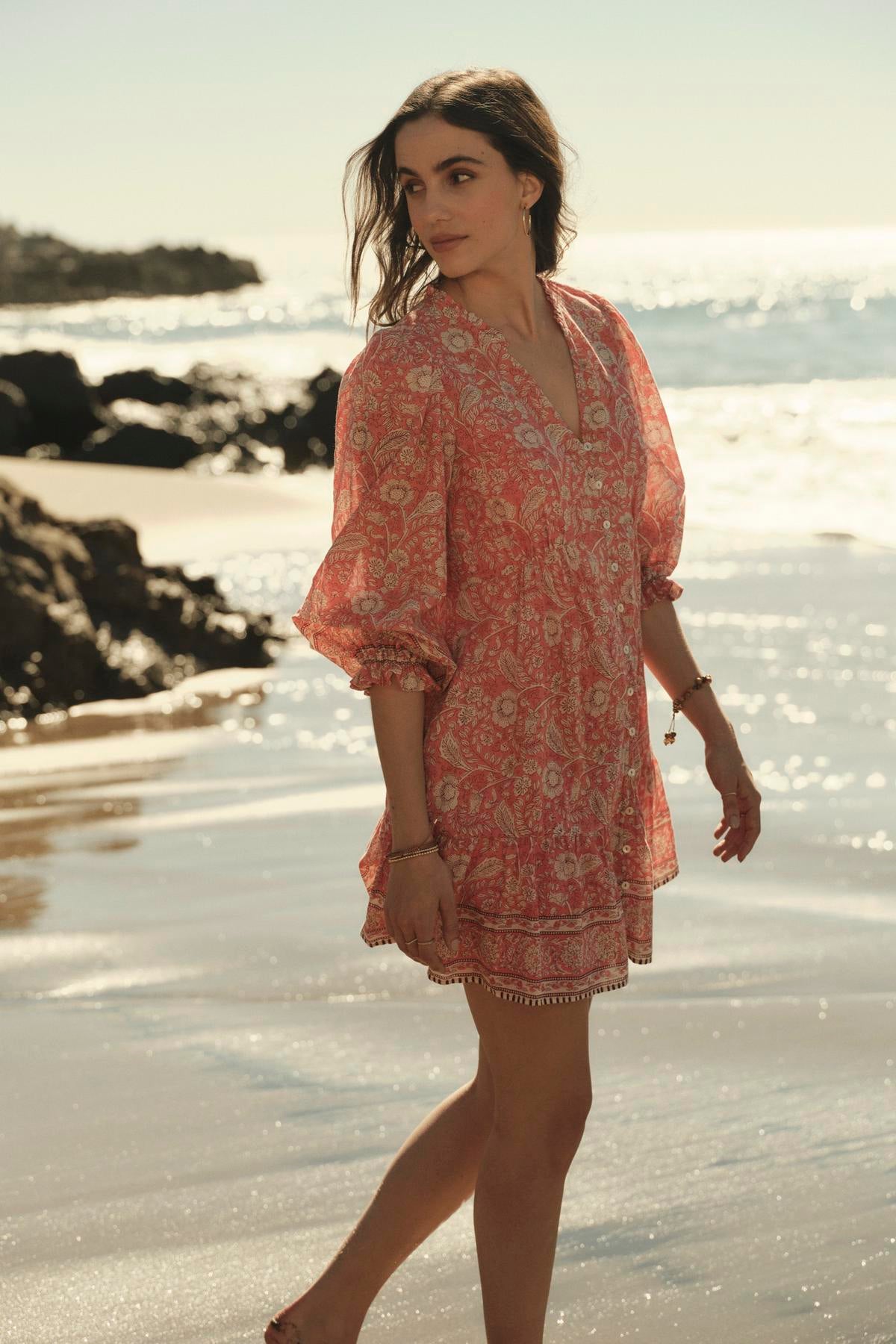 A woman in a Velvet by Graham & Spencer MARY DRESS walks along a beach at sunset, her gaze turned slightly away from the camera.-36910098907329