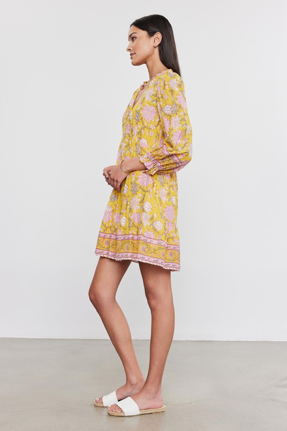   A woman stands sideways wearing the MARY DRESS from Velvet by Graham & Spencer, a yellow floral dress with pink accents, paired with white sandals against a plain background. The vibrant printed cotton voile dress showcases an airy, summery feel. 