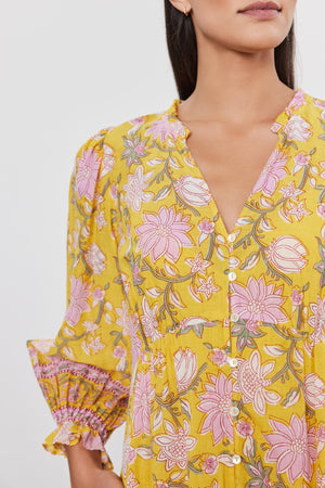 A woman wearing the MARY DRESS by Velvet by Graham & Spencer, a vibrant printed cotton voile dress in yellow with pink floral patterns and long sleeves. This tiered design dress features a button-down front and ruffled cuffs.