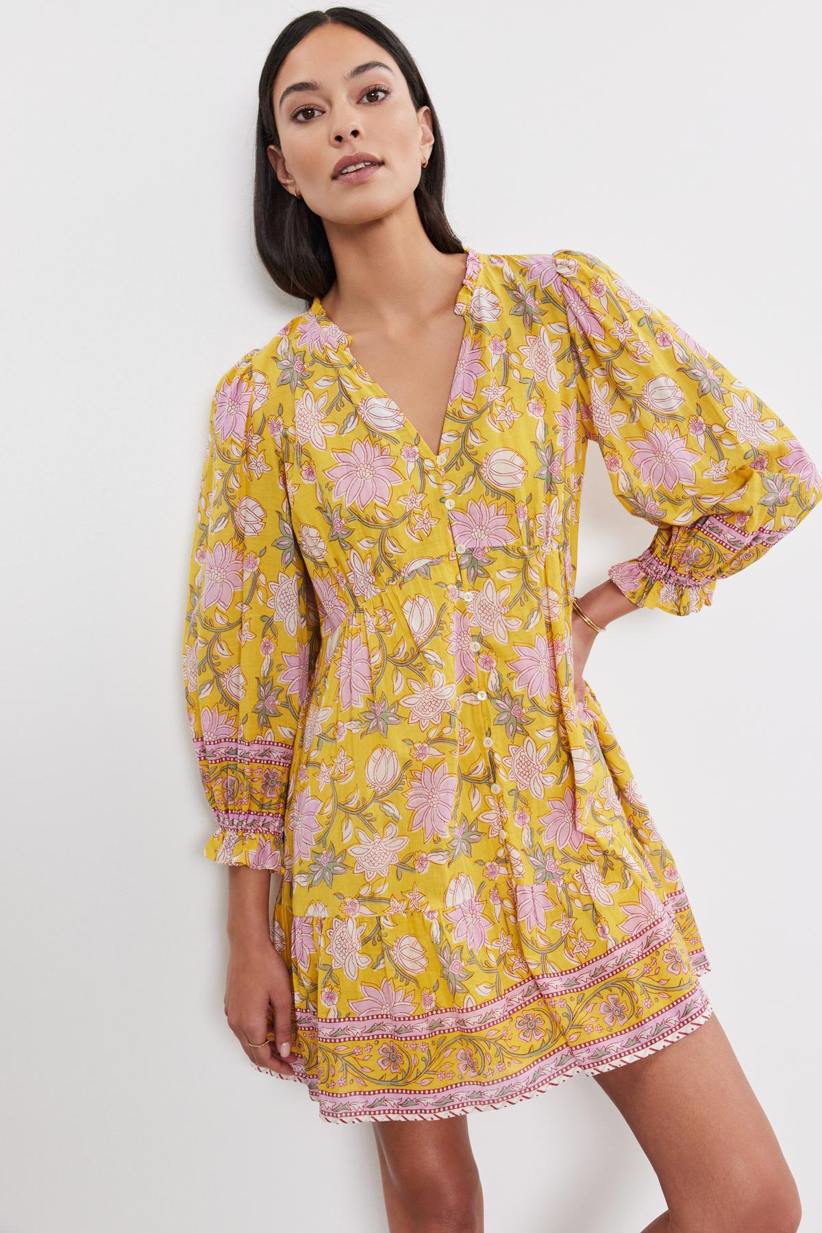   A woman in the Velvet by Graham & Spencer MARY DRESS, a yellow floral ensemble with long sleeves and a v-neckline, stands against a plain white background, showcasing its elegant tiered design. 