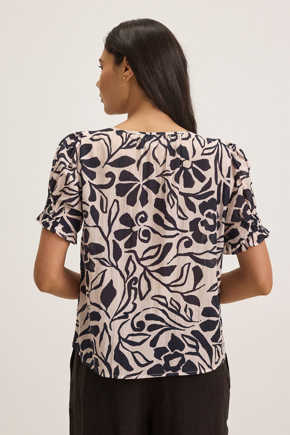   A woman with dark, long hair and a beige blouse featuring a black floral pattern is shown from behind. The top is the LESLIE TOP by Velvet by Graham & Spencer, set against a plain, light-colored background. 