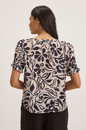 A woman with dark, long hair and a beige blouse featuring a black floral pattern is shown from behind. The top is the LESLIE TOP by Velvet by Graham & Spencer, set against a plain, light-colored background.