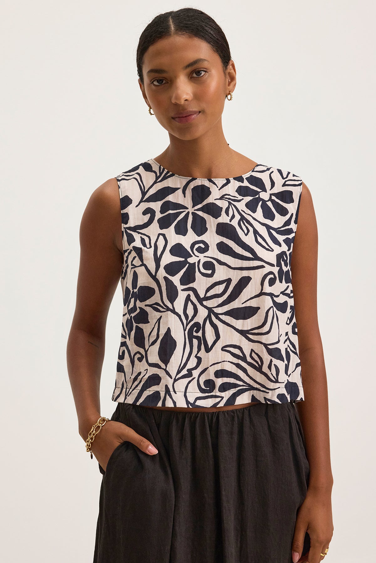 The person is wearing the NAEEM TOP by Velvet by Graham & Spencer, a cotton viscose sleeveless top in a black and white floral pattern, paired with a sleek black skirt. The outfit's cropped silhouette contrasts beautifully against the plain background for an effortlessly chic look.-38663151419585