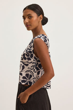 A person with dark hair in a bun wears the NAEEM TOP by Velvet by Graham & Spencer, a black and white floral cotton viscose sleeveless piece, paired with black pants against a plain background.