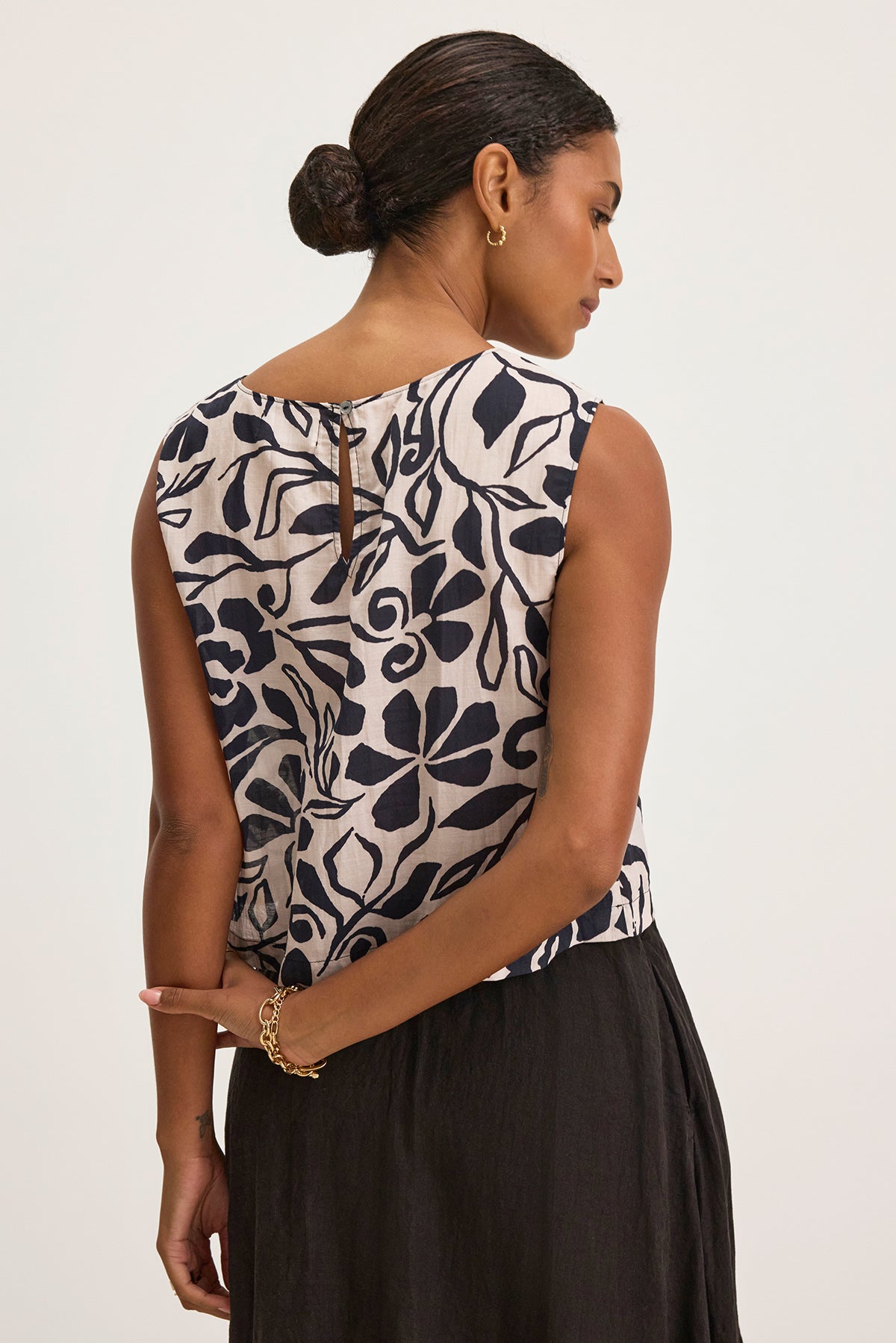   A woman with a bun wears the NAEEM TOP, a sleeveless cotton viscose top by Velvet by Graham & Spencer, featuring a navy leaf pattern. She pairs it with a black skirt and stands with her hands crossed behind her back. 