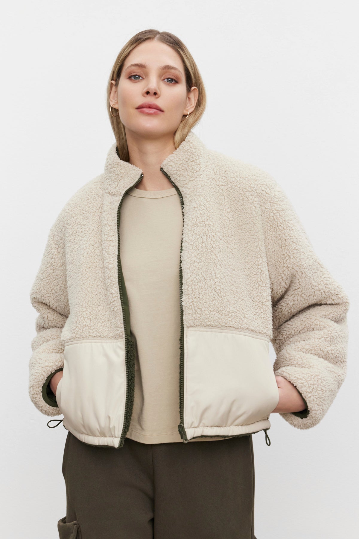   A person stands against a plain background, wearing the TASHA REVERSIBLE PUFFER SHERPA JACKET by Velvet by Graham & Spencer, featuring a high collar and hands in the front pockets. 