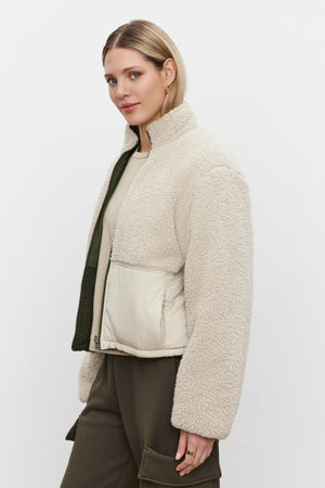 A woman with short blonde hair, dressed in a Velvet by Graham & Spencer TASHA REVERSIBLE PUFFER SHERPA JACKET over a light shirt and green cargo pants, is standing against a white background.