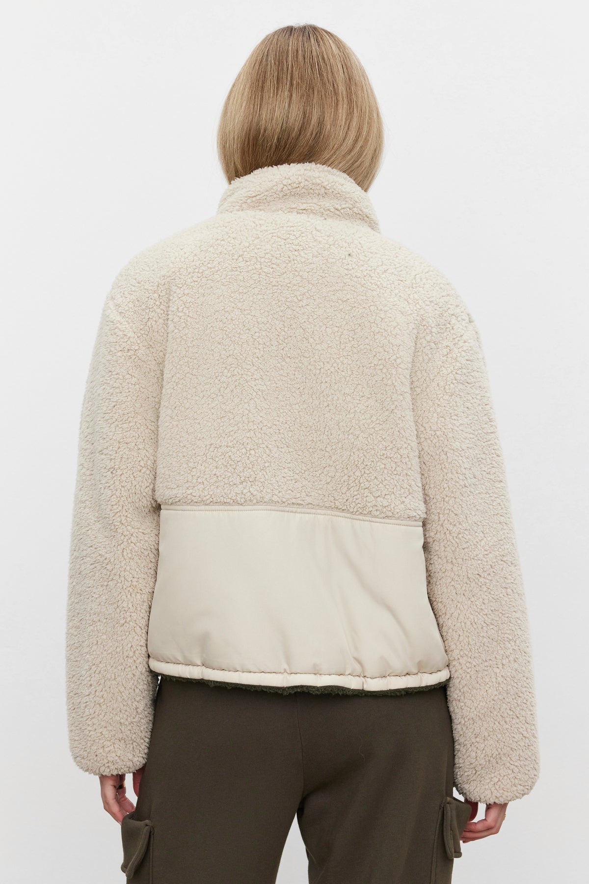   Woman seen from behind wearing the TASHA REVERSIBLE PUFFER SHERPA JACKET by Velvet by Graham & Spencer, featuring a beige faux sherpa design with a quilted section in the lower back, paired with olive green cargo pants. 