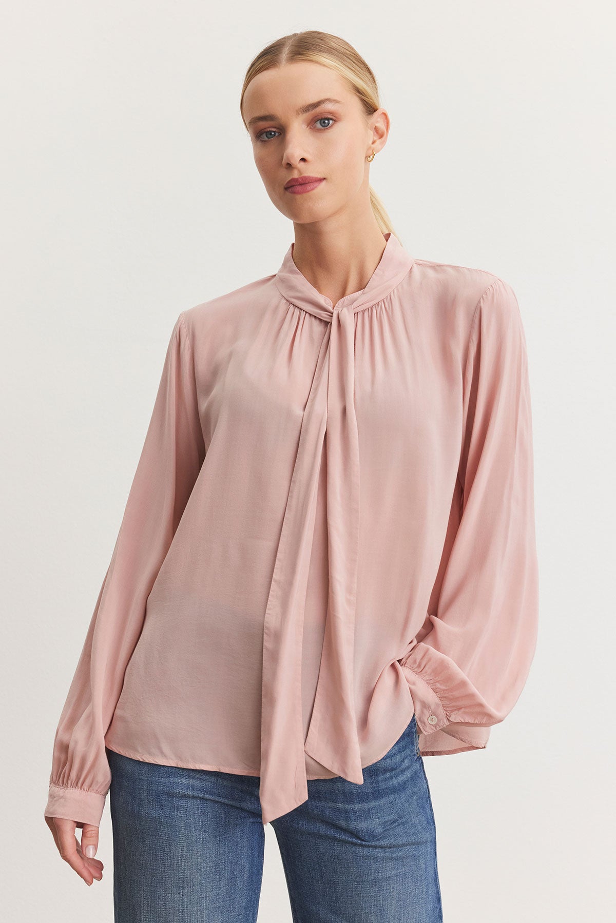   A person wearing the GABRIELLE TOP from Velvet by Graham & Spencer, a light pink rayon challis blouse with a tie-neck design, and blue jeans styled in a relaxed fit, stands against a plain background. 