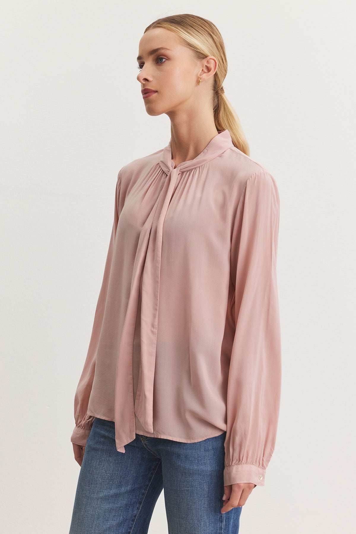   A woman wearing the GABRIELLE TOP in a relaxed fit light pink blouse with a banded collar from Velvet by Graham & Spencer, paired with blue jeans, stands against a plain white background. 