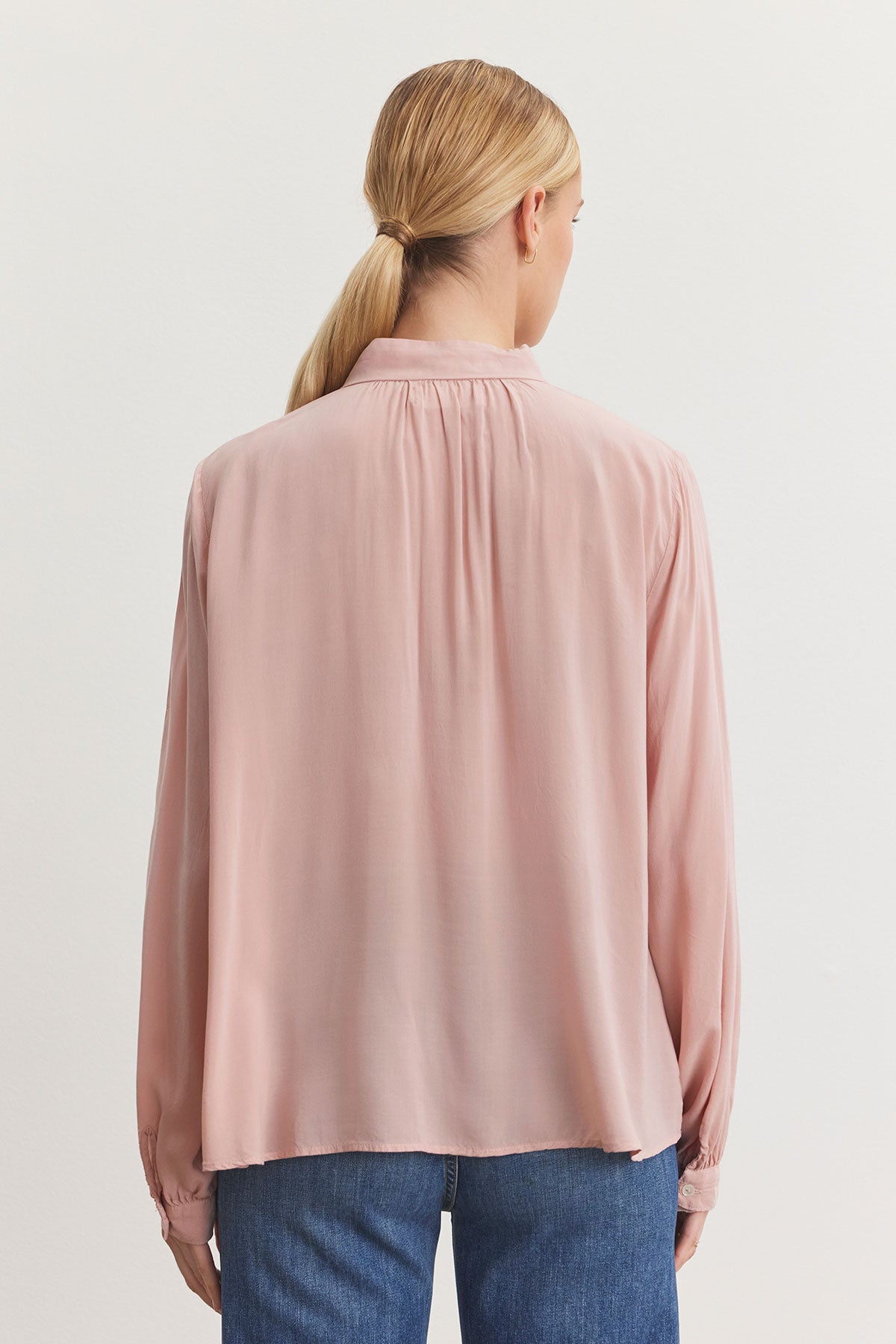   A person with long hair in a ponytail stands facing away, wearing the GABRIELLE TOP from Velvet by Graham & Spencer in a pink hue. This blouse is crafted from rayon challis fabric and paired with blue jeans, its relaxed fit beautifully complementing the banded collar for a touch of casual elegance. 