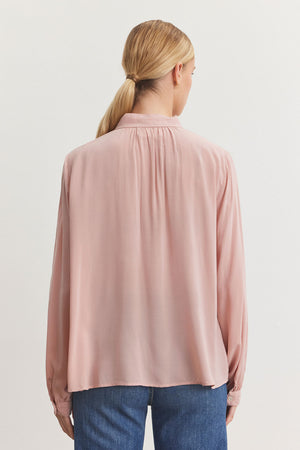 A person with long hair in a ponytail stands facing away, wearing the GABRIELLE TOP from Velvet by Graham & Spencer in a pink hue. This blouse is crafted from rayon challis fabric and paired with blue jeans, its relaxed fit beautifully complementing the banded collar for a touch of casual elegance.